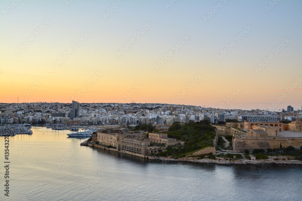 The photograph presents a panoramic view of a harbor in Malta at dusk, featuring a cityscape with historic buildings and fortisifications along the waterfront
The photograph presents a panoramic view of a harbor in Malta at dusk, featuring a cityscape with historic buildings and fortisifications along the waterfront
