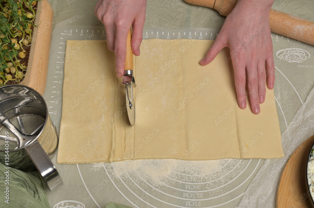 Woman's hands cuts a sheet of dough. Rolling pin, cutter, green towel, microgreens sprouts on kitchen table. Home cooking baking puffs Woman's hands cuts a sheet of dough. Rolling pin, cutter, green towel, microgreens sprouts on kitchen table. Home cooking baking puffs