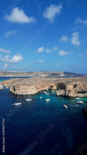 Vertical aerial drone view of the Blue Lagoon on Comino Island, Malta. Panorama of the Mediterranean Sea, yachts, boats, and people standing on stunning cliffs.