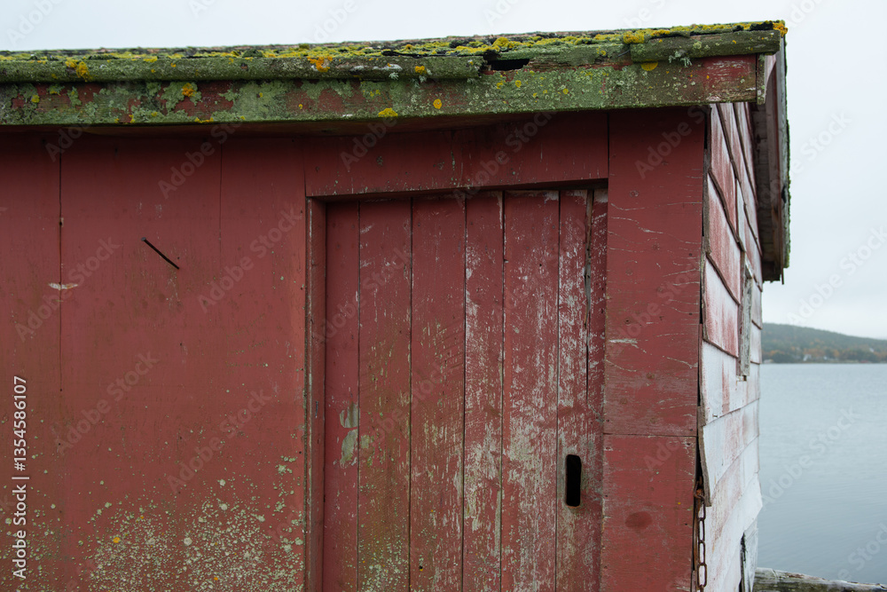 A worn and weathered red wooden fishing shed. There's a single solid door with a black metal handle. The roof is damaged and covered with green moss and lichen. The wall is clapboard wood siding. A worn and weathered red wooden fishing shed. There's a single solid door with a black metal handle. The roof is damaged and covered with green moss and lichen. The wall is clapboard wood siding.