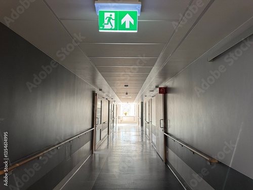 Long and empty hospital corridor with bright lights from window and green evacuation exit sign in the foreground