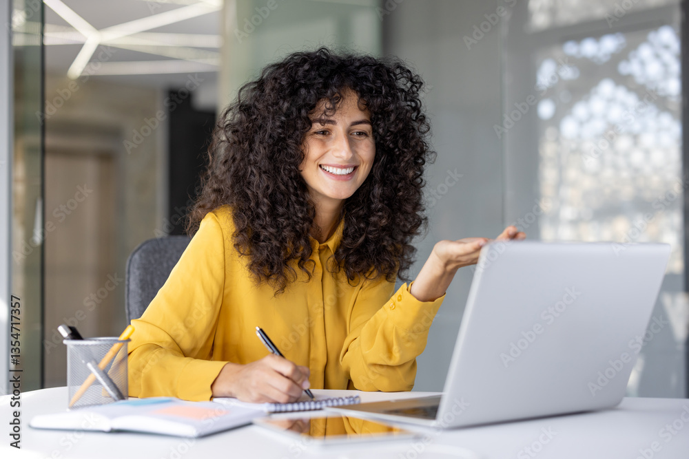 Smiling woman in yellow shirt is attending an online course, taking notes while gesturing in front of her laptop