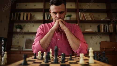 A man in pink shirt is deeply focused on chess game, with his hands clasped together in front of him,analyzing the board with black and white pieces. Bookshelves and lamp are visible in the background