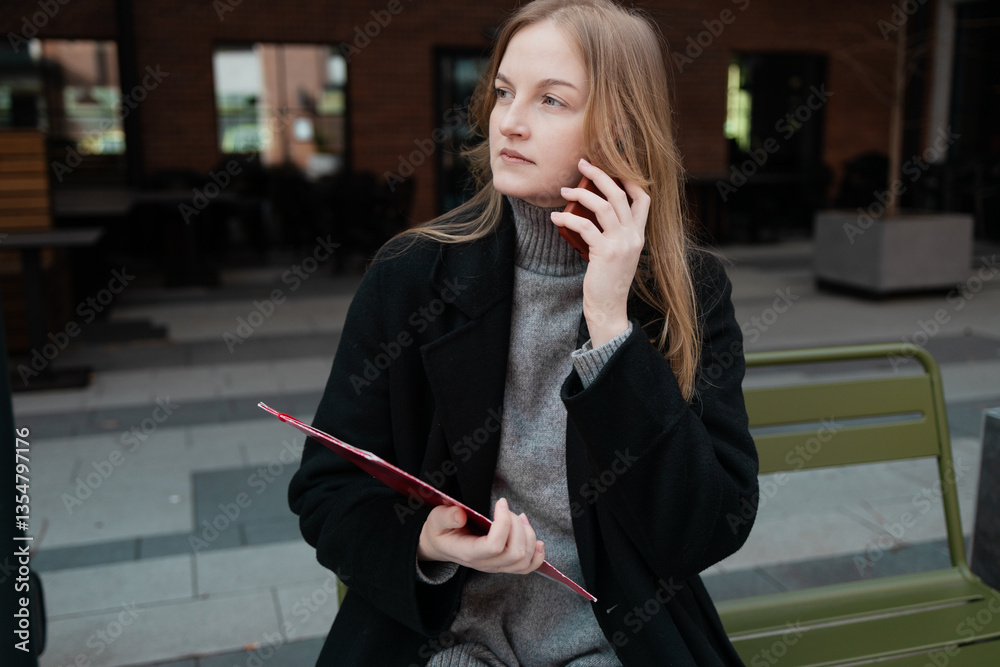 Business woman with a red folder with documents talking on mobile phone outdoors, professional woman in suit sits on a bench near the business center