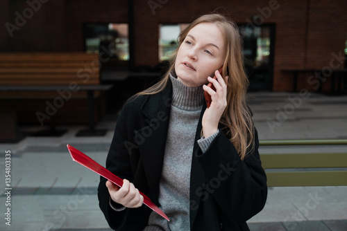 Obraz na płótnie Business woman with a red folder with documents talking on mobile phone outdoors, professional woman in suit sits on a bench near the business center w salonie