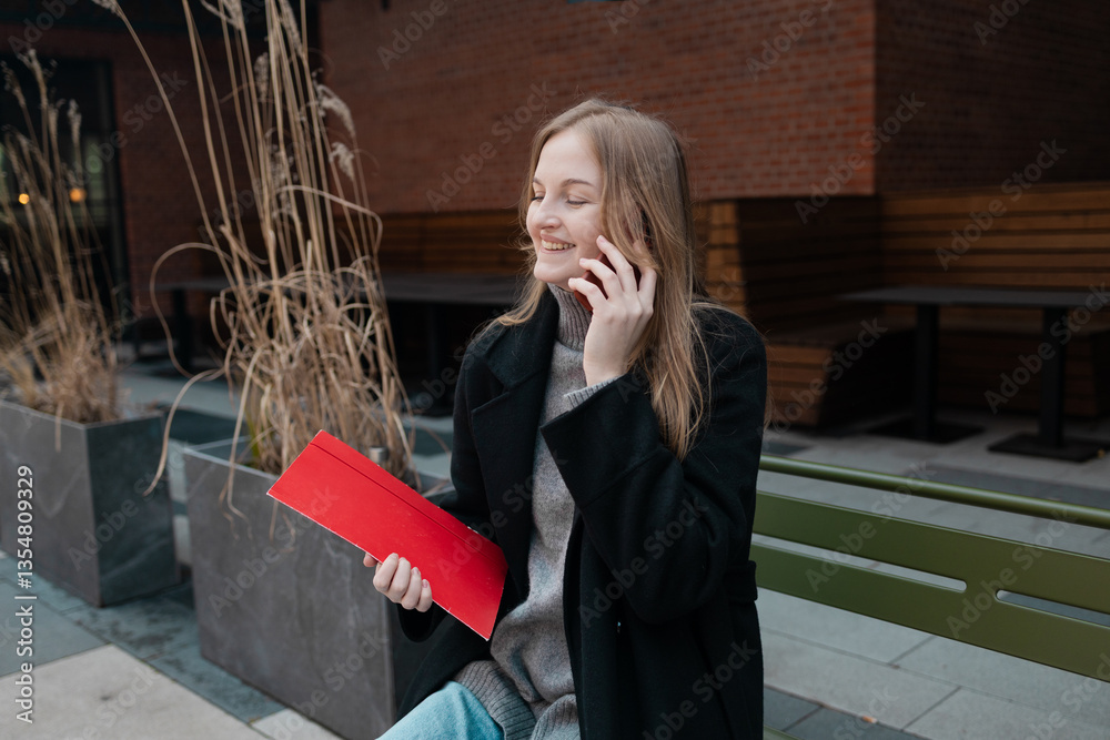 Business woman with a red folder with documents talking on mobile phone outdoors, professional woman in suit sits on a bench near the business center