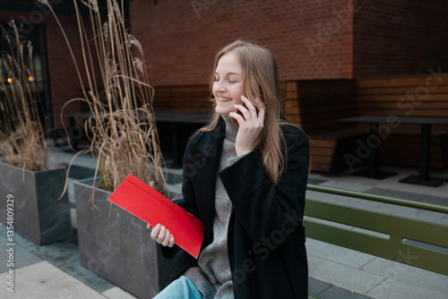 Obraz na płótnie Business woman with a red folder with documents talking on mobile phone outdoors, professional woman in suit sits on a bench near the business center w salonie