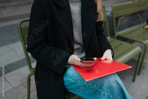 Obraz na płótnie Photo of successful confident business lady holding folder with documents while sitting on street outdoors against the background of the business center. A woman looks at her phone and checks the news w salonie