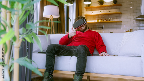 Obraz na plátně Young man in a red sweater immersed in a VR first-person shooter game, sitting on a cozy white couch in a modern living room, experiencing an intense moment while switching weapons in virtual reality