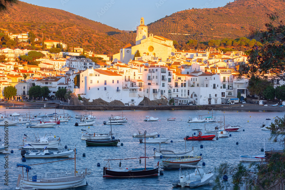 View of fishing boats in the bay of Cadaques in Catalonia, Spain with white buildings and church at sunset