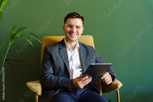 Professional man sits comfortably in a modern chair holding a tablet in a stylish green office space photo