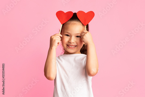 Love Concept. Playful asian girl holding two paper hearts over pink studio background, copyspace