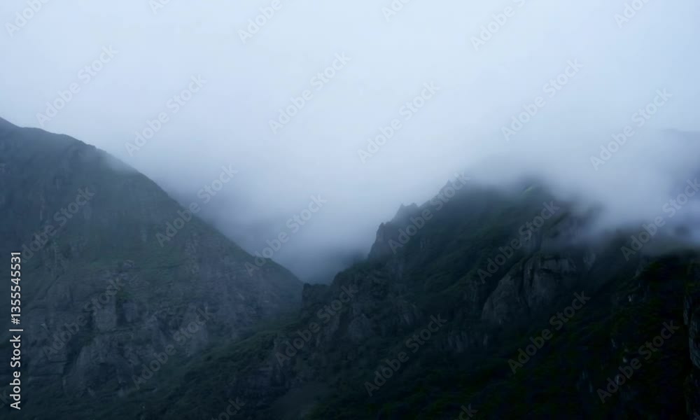 Thick fog drifts slowly over dark, rugged mountain peaks, partially obscuring the landscape. The light is diffused, casting a moody, cinematic atmosphere. The scene feels vast and mysterious