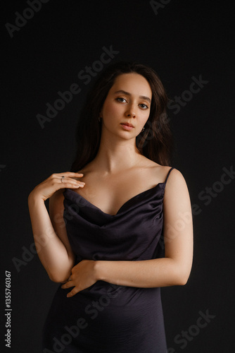 Caucasian woman in black satin dress on dark background. Touching shoulder, front view, looking into camera. Concepts of beauty, skincare, cosmetics, self-care, and luxury fashion.