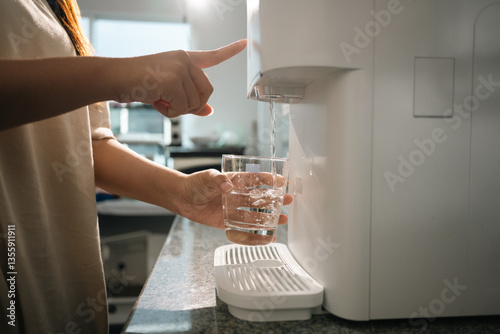 Woman using a modern water purifier in kitchen, filling a glass with fresh, purified water