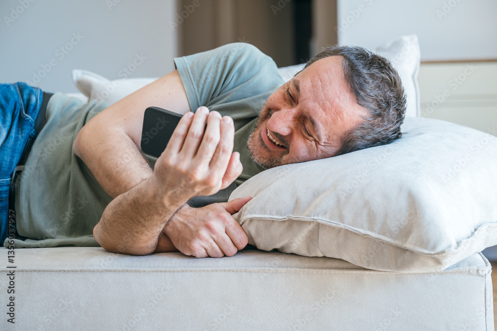 Casual male watching entertaining social media content on mobile phone device while laying on living room sofa Casual male watching entertaining social media content on mobile phone device while laying on living room sofa