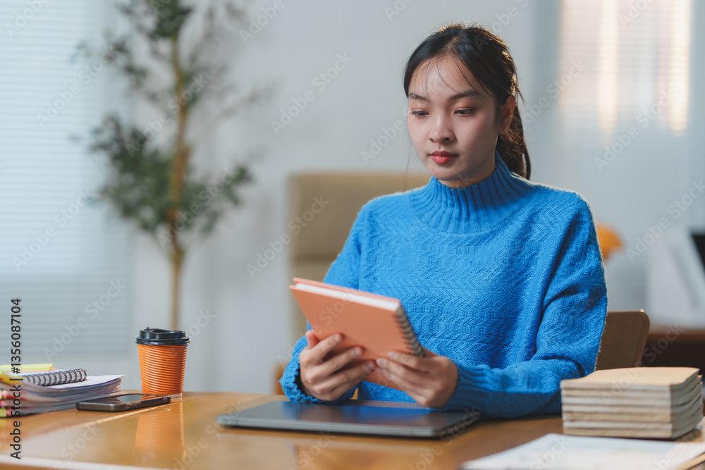 Fototapeta premium Young woman in a blue sweater reading a notebook at a desk in a modern office. Coffee cup and documents surrounding her suggest a focused work environment