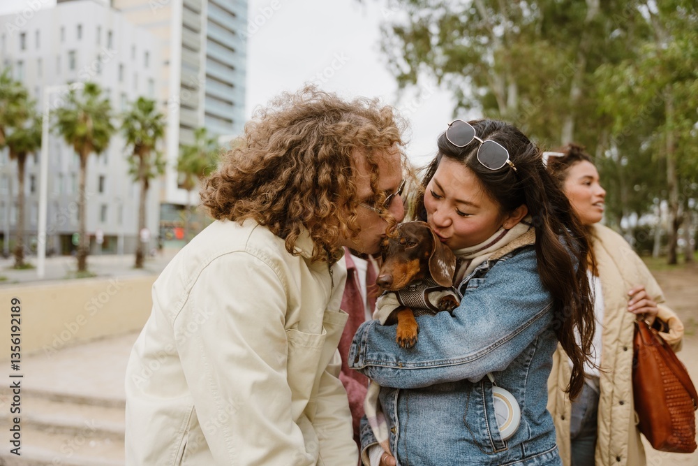 Obraz premium An adult Asian woman and an adult White man wearing sunglasses hold a dachshund on a street with buildings in the background. They are kissing the dog. An adult Latino woman stands in the background.