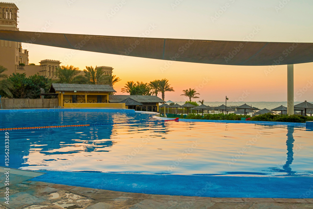 Luxury hotel pool in Sharjah at sunset, with golden light reflecting on the water. Elegant sun loungers line the poolside, offering a serene view of the beach and tranquil sea waves.