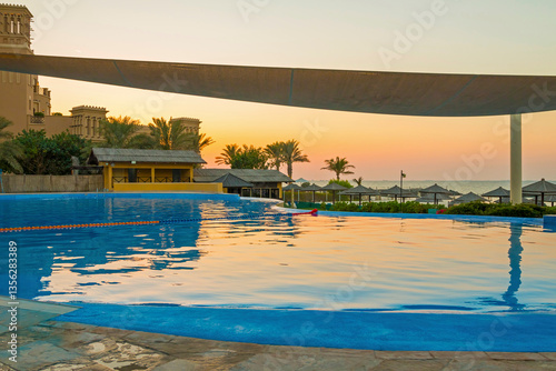 Obraz na płótnie Luxury hotel pool in Sharjah at sunset, with golden light reflecting on the water. Elegant sun loungers line the poolside, offering a serene view of the beach and tranquil sea waves. w salonie