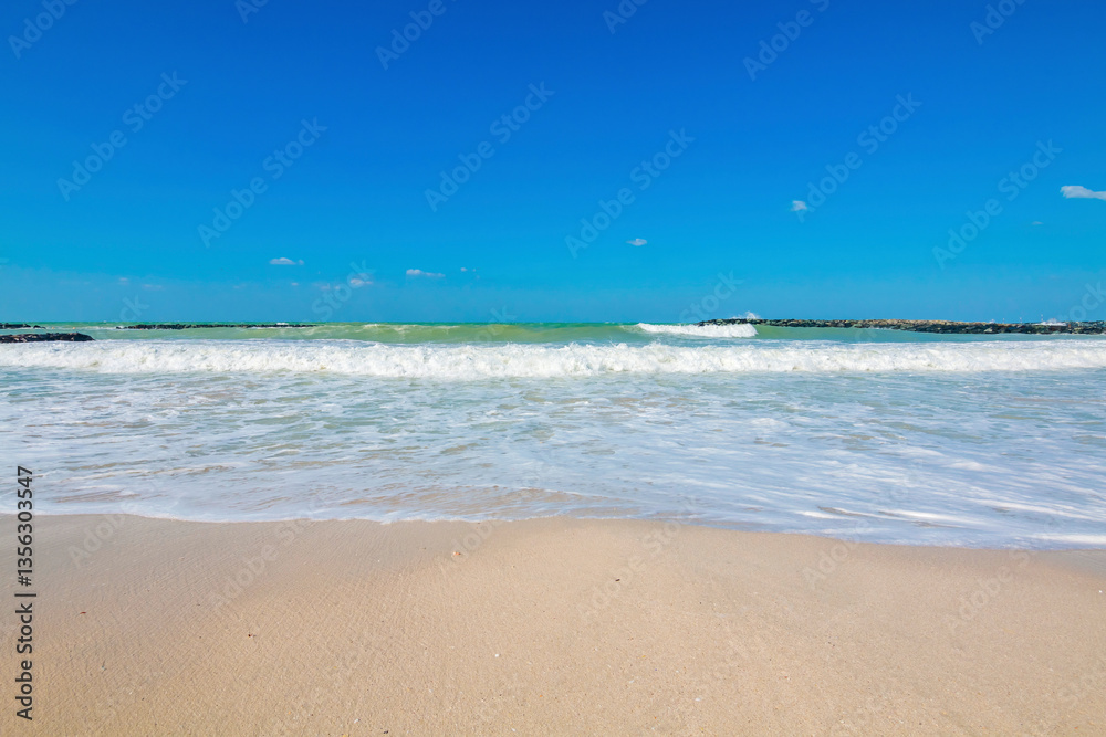 Pristine beach in Sharjah on a sunny day, with golden sand stretching along the shoreline. The calm turquoise waters gently lap against the shore, under a clear, cloudless blue sky.
