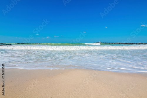 Obraz na płótnie Pristine beach in Sharjah on a sunny day, with golden sand stretching along the shoreline. The calm turquoise waters gently lap against the shore, under a clear, cloudless blue sky. w salonie