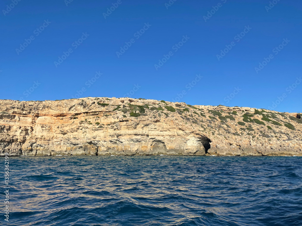 A towering coastal cliff with distinct rock layers rising above the deep blue sea under a clear sky