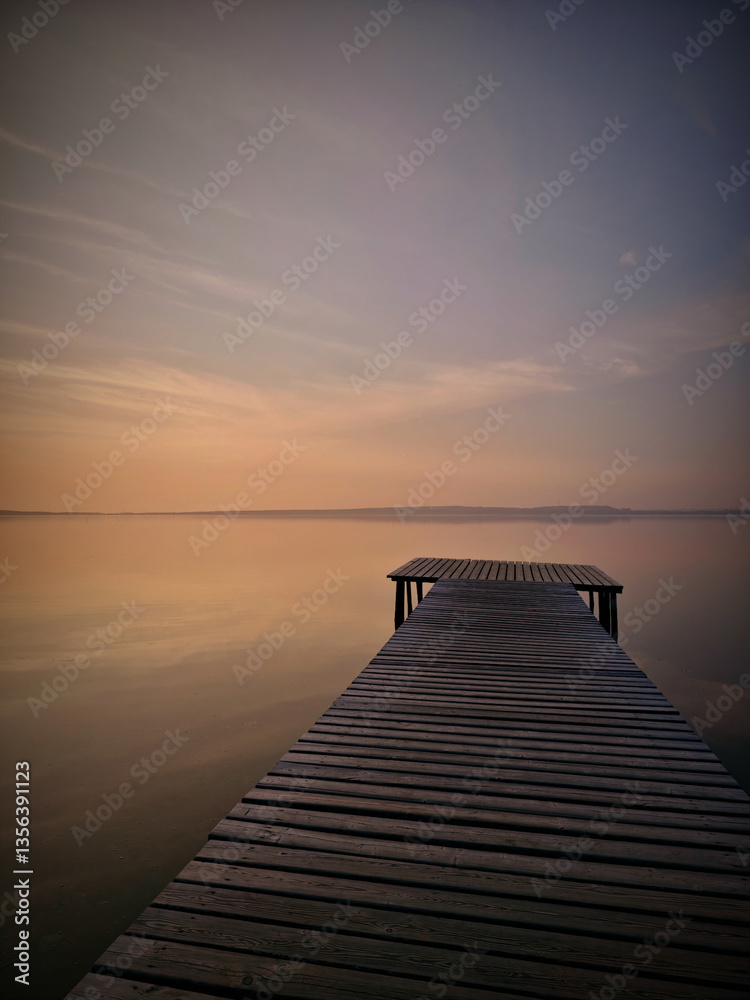 Obraz premium A serene view of a wooden dock extending into a vast body of calm water at sunset. the sky is painted in soft hues of orange and pink, creating a peaceful ambiance. the tranquil setting evokes a sense