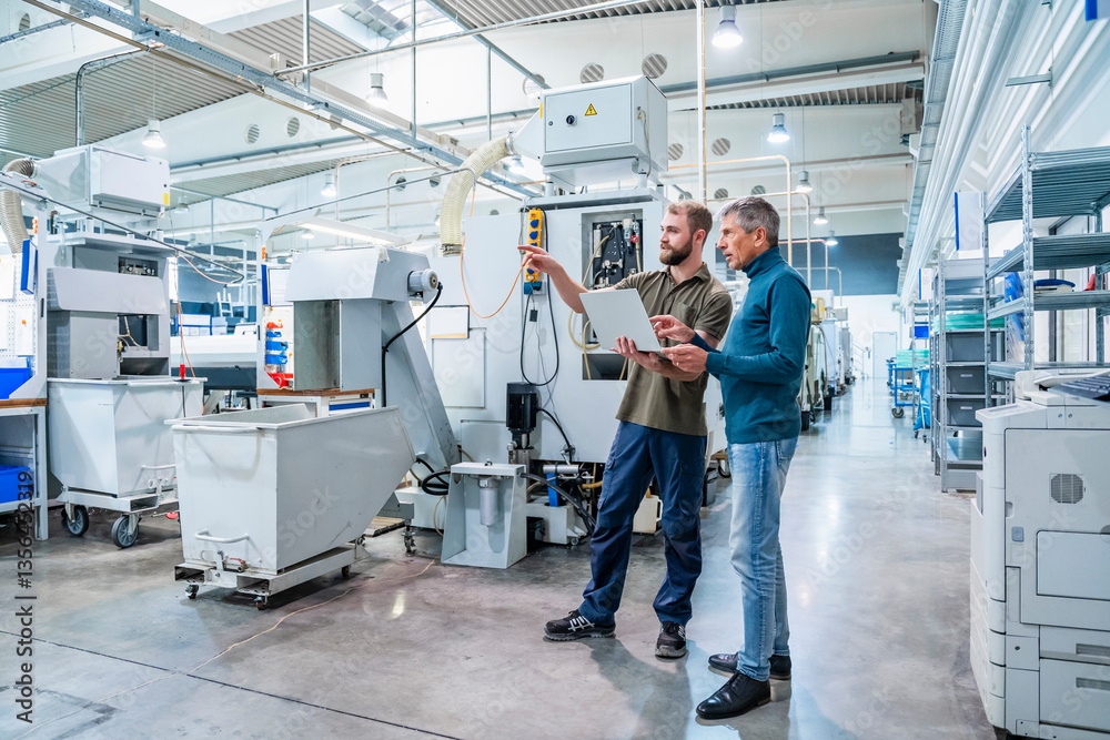 Two men discussing in a production hall with machinery and a tablet Two men discussing in a production hall with machinery and a tablet