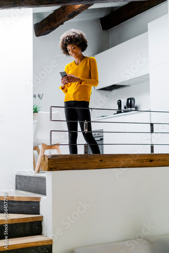 Smiling young woman standing in the kitchen using smartphone