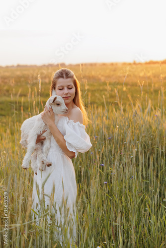 A young woman in a white dress stands in a field at sunset, gently holding a white baby goat. The warm evening light and golden landscape create a peaceful, idyllic atmosphere of nature and harmony.