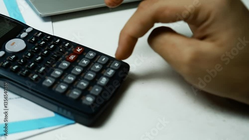 Person Using Scientific Calculator at Desk, A person using a scientific calculator with a laptop in the background