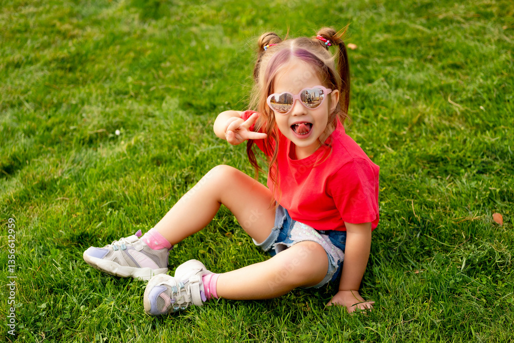 Fototapeta premium a baby girl on a green lawn, a happy little child in glasses and a red T-shirt lying on the grass in summer, a place for text