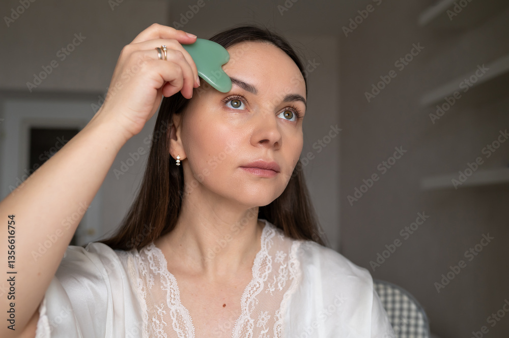 Young woman in silk robe doing facial massage with gua sha scraper while looking in mirror. Home skin care and aesthetic procedures Young woman in silk robe doing facial massage with gua sha scraper while looking in mirror. Home skin care and aesthetic procedures