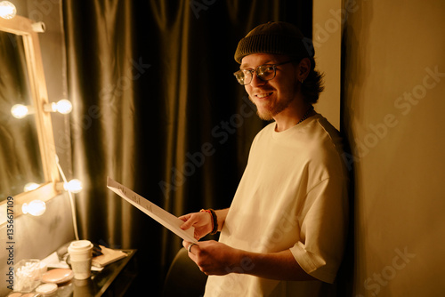 Young man wearing beanie reading script in dimly lit dressing room with vanity lights creating warm ambiance