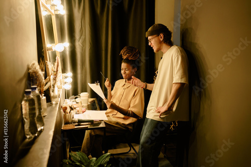 Two actors preparing backstage in theater dressing room with bright lighting and makeup items on vanity table Modern atmosphere with collaborative engagement and focused expressions