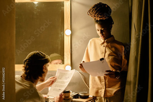 Team of filmmakers reviewing script in backstage area with thoughtful expressions, preparing for upcoming scenes. Lighting coming from surrounding lamps illuminating faces and workspace