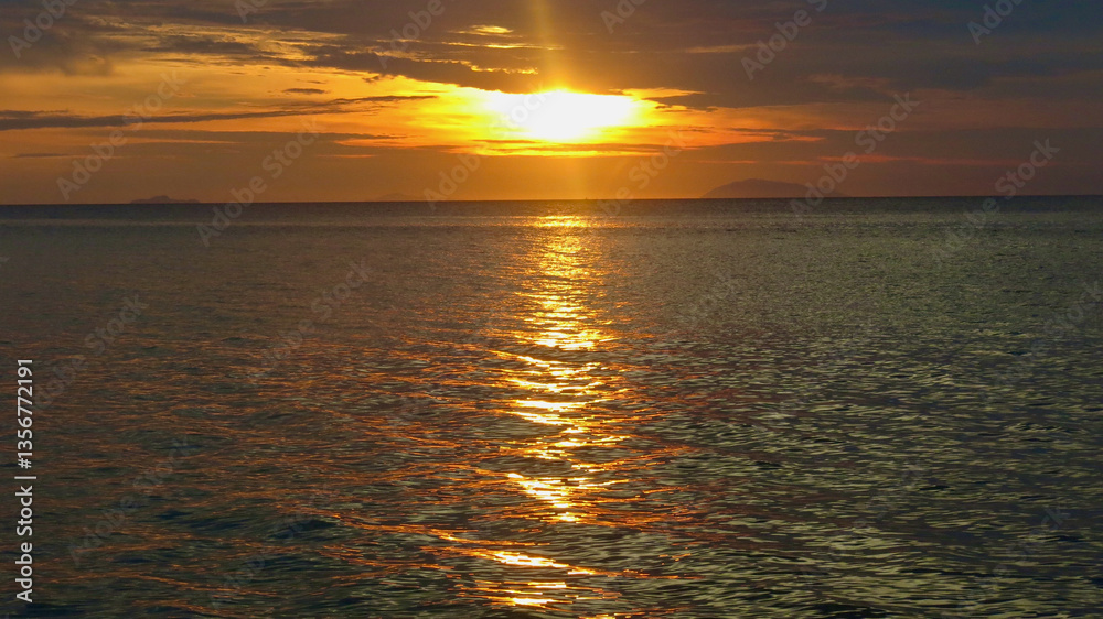 Fototapeta premium Sunset over the sea. View of the calm surface of the ocean during sunset. The sun sets behind the sea behind the clouds.