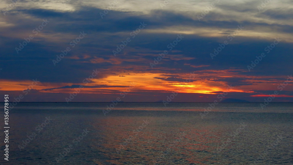Obraz premium Sunset over the sea. View of the calm surface of the ocean during sunset. The sun sets behind the sea behind the clouds.