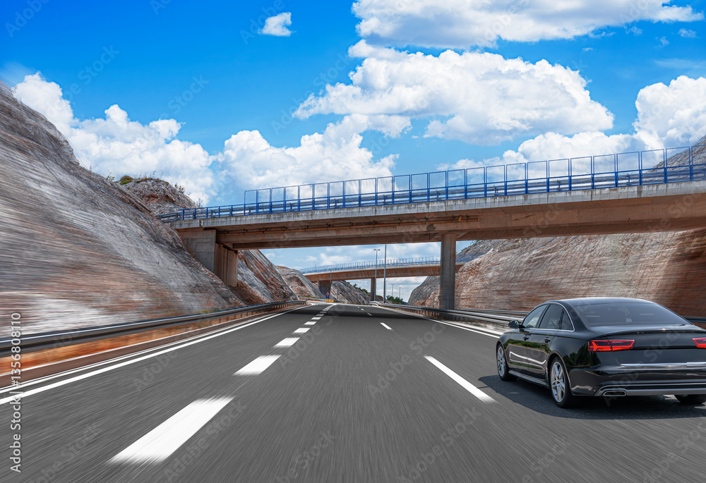 A black luxury sedan speeds along a modern highway through a mountainous landscape, passing under bridges with railings beneath a bright blue sky with scattered clouds.