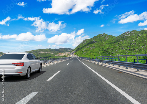 A white sedan drives along a modern highway surrounded by lush green mountains, under a bright blue sky with scattered clouds, creating a scenic and peaceful road trip atmosphere.