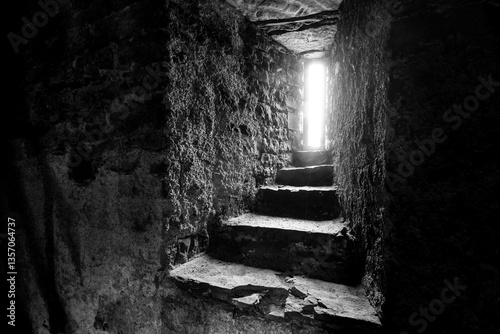 Small opening in a very thick quarry stone wall in the steeple of a centuries-old fortified church in Germany. High contrast black and white image with incidence of light and stairway steps.