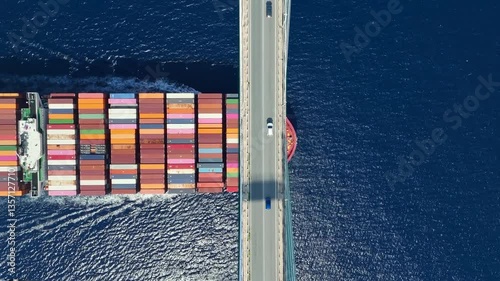 Transport concept with an aerial overhead view of a very big container super cargo ship passing under a bridge with car traffic