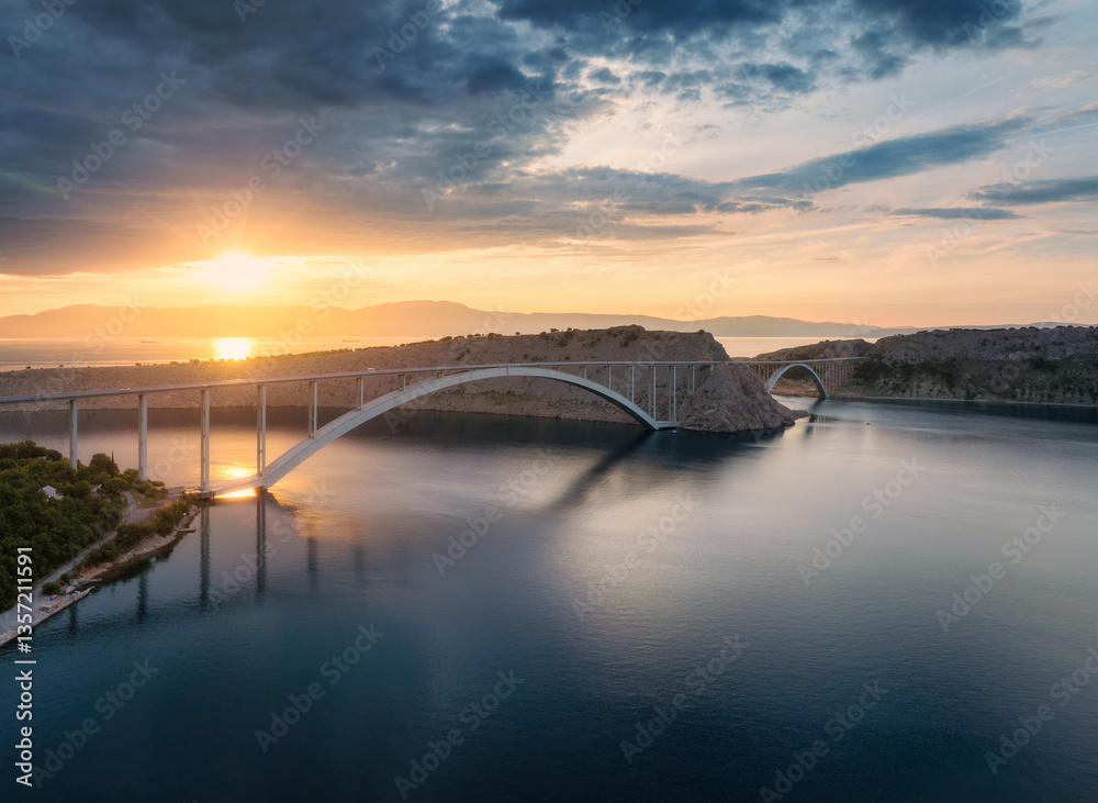Naklejka premium Aerial view of bridge and blue sea at colorful sunset. Modern Krk bridge island during golden hour, mountains, rocks, reflection in water and cloudy sky. Top drone view of bridges. Architecture