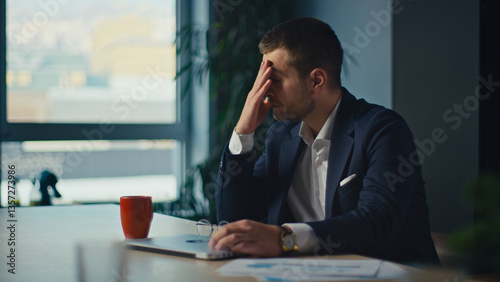 Stressed businessman in a suit sits at a desk, rubbing his forehead in frustration. A laptop, documents, and a coffee cup are on the table. Concept of workplace stress and business challenges