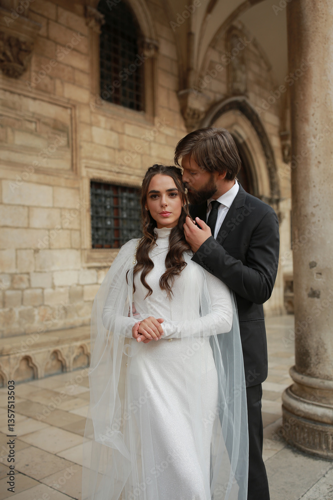 A wedding couple poses near a majestic historic building in Croatia. The bride, in a luxurious white gown with a long train, and the groom, in a classic suit, create an atmosphere of romance A wedding couple poses near a majestic historic building in Croatia. The bride, in a luxurious white gown with a long train, and the groom, in a classic suit, create an atmosphere of romance