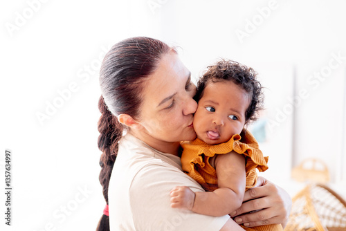 a white-skinned mother hugs and kisses an African-American black baby girl at home by the window, maternal love and care, a mother of European appearance with a black child in her arms