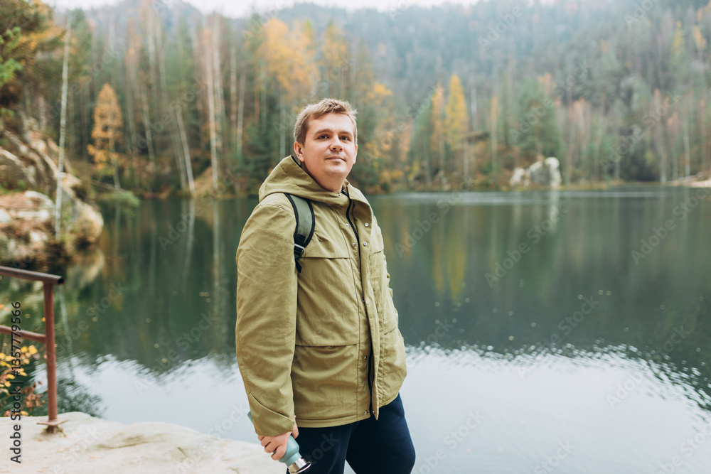 A smiling 30s blonde male traveler with a backpack enjoys the beautiful scenery of the majestic lake. Travel, adventure. Concept of an active lifestyle.