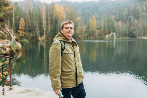 Obraz na płótnie A smiling 30s blonde male traveler with a backpack enjoys the beautiful scenery of the majestic lake. Travel, adventure. Concept of an active lifestyle. w salonie