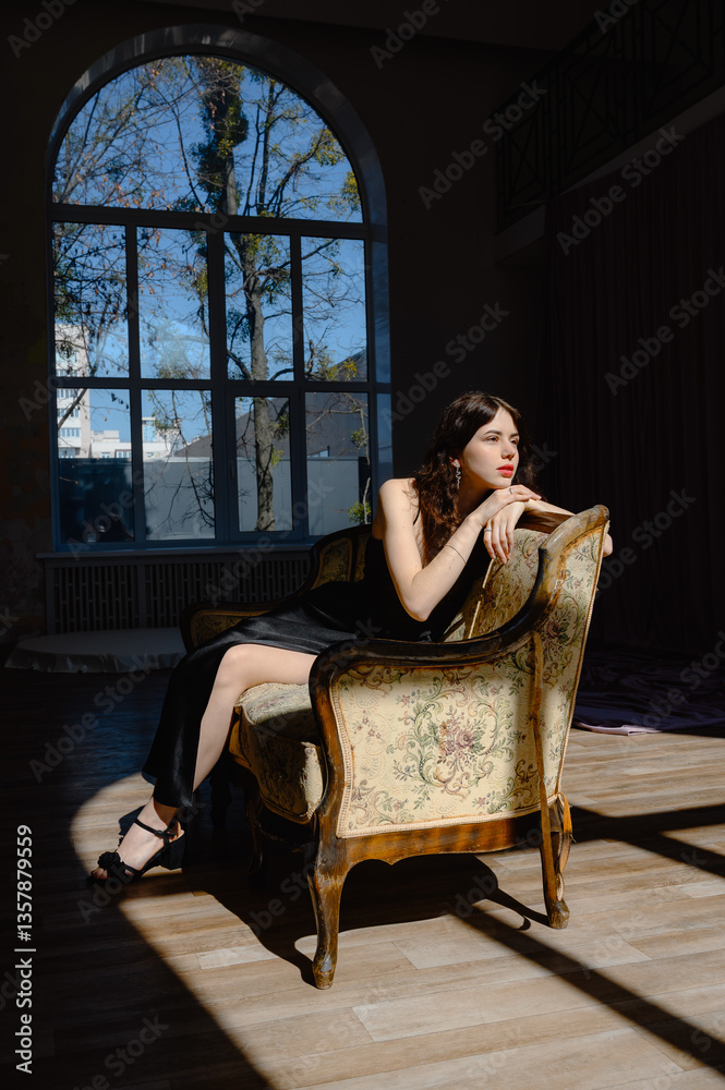 Obraz premium An elegant woman in a black dress sits on a sofa. in the background a large window style and beauty Young girl